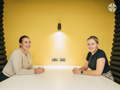 2 people sitting across a white table having a friendly conversation in a meeting pod with yellow walls and acoustic panels.