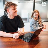 Two colleagues, a white man and a white woman, laugh together over laptops at a meeting table in a modern office.