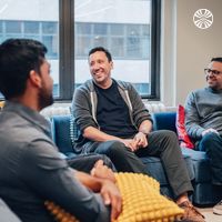 Four team members engaged in a casual conversation while seated on comfortable lounge furniture in a bright office space.