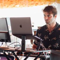 A white man focused on working at a standing desk with a laptop and monitor, in a brightly lit office space with an orange ceiling.