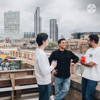 A racially diverse group of 3 men chatting and holding cups on a rooftop, with a cityscape in the background featuring tall buildings.