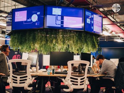 Modern workspace featuring hanging plants and digital displays, with several employees working at ergonomic desks.