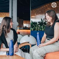 2 white women seated and laughing indoors, each holding a Jigsaw branded bottle.