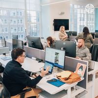 Team members in smart casual clothes work at individual desks in an open-plan office. The office has wooden floors and natural lighting.