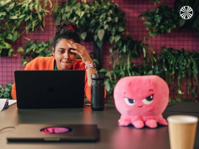 Employee working next to a pink octopus plush mascot displayed near office greenery and computer screen.