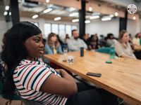 A Black woman listens intently during a company meeting with diverse colleagues seated around her in the background.