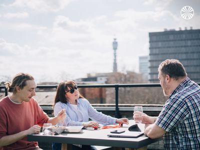 Three team members having a rooftop lunch meeting with London skyline and communication tower in background.