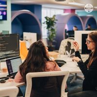 Two colleagues in conversation at their workstations in a modern office space with arched architectural details.