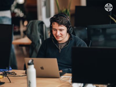 White man smiling, working at his laptop with headphones in a relaxed office space.