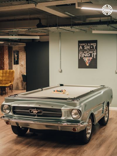 Classic silver Mustang car shaped pool table in an office recreation area with motivational poster on wall.