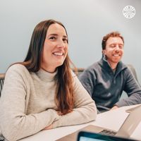 Two team members smiling at the camera. They're seated with laptops in a meeting room. 