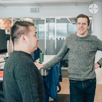 3 people standing engaged in casual conversation in an open office space with glass partitions.