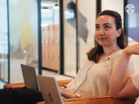 A woman with medium-length dark hair sits with a laptop in a modern, open-office environment, looking attentive.