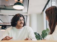 Two people in casual attire sit at a table, engaged in a discussion in a brightly lit office space.