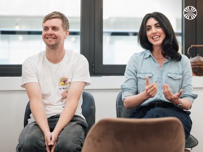 An animated white woman gestures while presenting during company all hands. Her white male colleague sits beside her smiling.