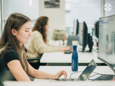 Person focused on laptop work at desk with water bottle, another colleague visible in background.