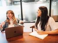 A white woman and a British Indian woman share a joyful moment in a meeting. The white woman is laughing and gesticulating. 