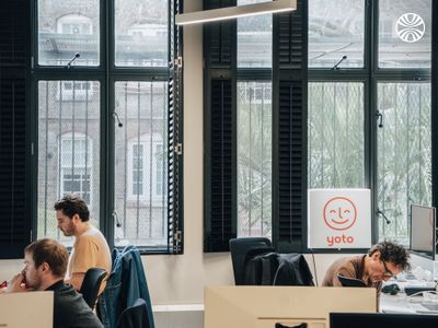 Three employees working at individual desks in bright office space with tall windows and Yoto branding.