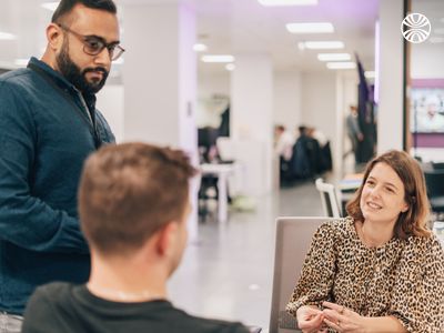 Three colleagues having a friendly discussion around a small table in a bright, open office setting.