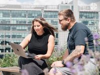 Two people sit outside with a laptop, discussing work. Modern building in the background.