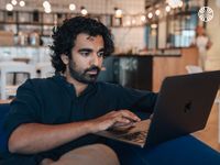 A focused man with curly hair uses a laptop, sitting in a comfortable, open-office space with a kitchen and shelves behind him.