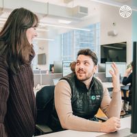 A person stands besides their colleague's desk. The seated team member is turned towards them in conversation. They are wearing a Dataiku-branded gilet.