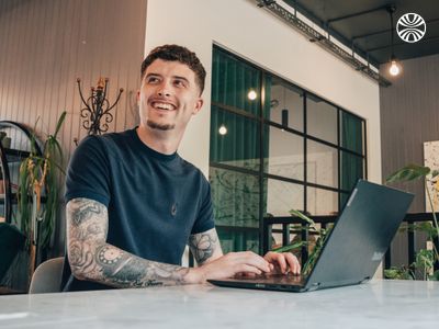 White man with tattoos smiling while working on his laptop at a shared desk.