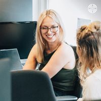 Two white women in a conversation at their desks, one smiling.