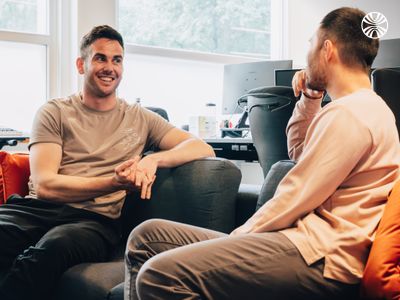 White male colleagues chatting on a couch in a relaxed workspace.