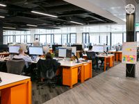 A large open workspace featuring orange desk units, ergonomic chairs, and a collaborative whiteboard area.