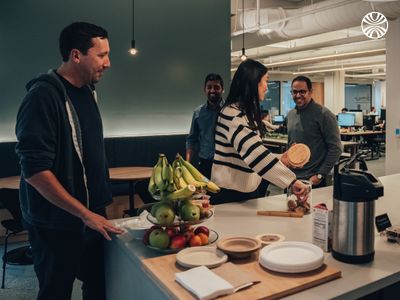 Four colleagues gathered around office kitchen counter with fresh fruit and coffee supplies during break time.