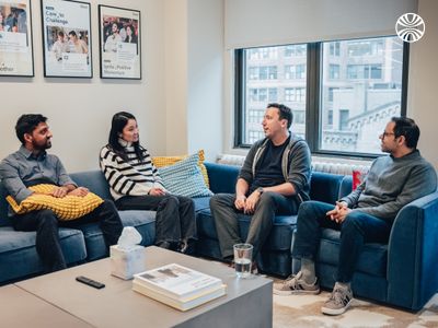 Four team members having an informal discussion while seated on blue couches, with framed artwork and city views in the background.