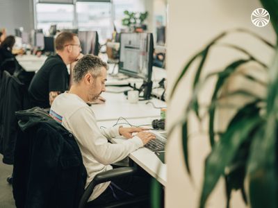 Person working at computer on a bank of desks in an open office space, with plants and colleagues visible in the background.