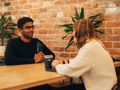 2 people having a friendly conversation over coffee at a wooden table against an exposed brick wall.