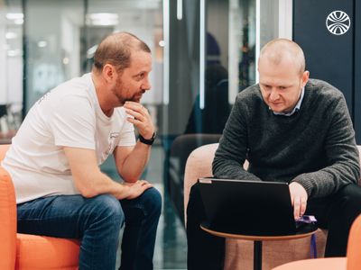 Two colleagues collaborating on a laptop while seated on orange chairs in a modern meeting space.