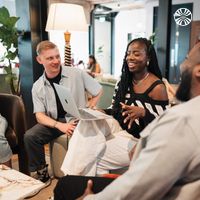 A racially diverse group of 4 colleagues engaged in a lively discussion while seated with laptops in a modern office lounge.