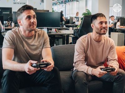 Two white male colleagues playing video games on a couch in the office.