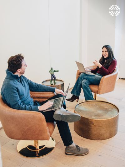 Two colleagues engaging in conversation. They are sitting on tan leather chairs facing each other with laptops on their laps.