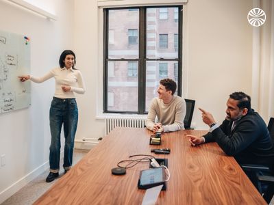 Three team members in a meeting room, with one person standing and presenting ideas on a whiteboard while colleagues listen attentively.