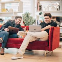 Two colleagues working on laptops and sharing a casual moment on a red velvet couch, 