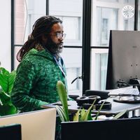 Colleague working at a desk in front of large black-framed windows with plants and natural light.