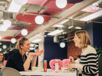 Two colleagues sharing lunch and conversation at a casual office dining area.