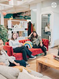 Team members sitting comfortably on a red velvet couch with a rainbow pillow in a casual break area. There are bookshelves and a phone booth behind them.