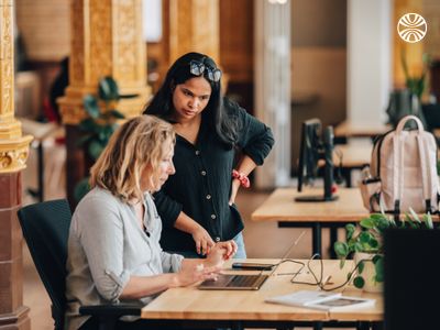 South Asian woman standing beside a white colleague seated at a desk, pointing at the screen as they work together.