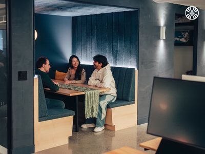 Four team members standing casually in a bright office space with modern furniture and audio-visual equipment visible in background.