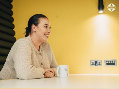 Person in a cream sweater smiling warmly during a conversation, leaning on a table against a yellow wall.