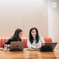 Two colleagues sitting in a red booth and working side by side on laptops.