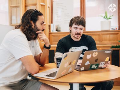 Two white men working on laptops at a table, focused on their screens, in a casual workspace.