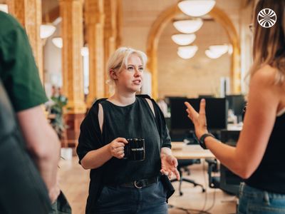 White woman with short blonde hair holding a mug, listening attentively to a colleague in a workspace.