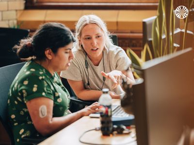 South Asian woman and white woman working together at a computer, engaged in discussion in an office.